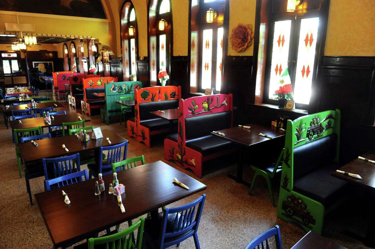A dining room of a restaurant with dark wood tables with napkin-wrapped silverware and bottles of salsa. Chairs and booth seating in various colors of blue, green, red and orange. The walls are yellow, and there are four stained glass windows to the right.