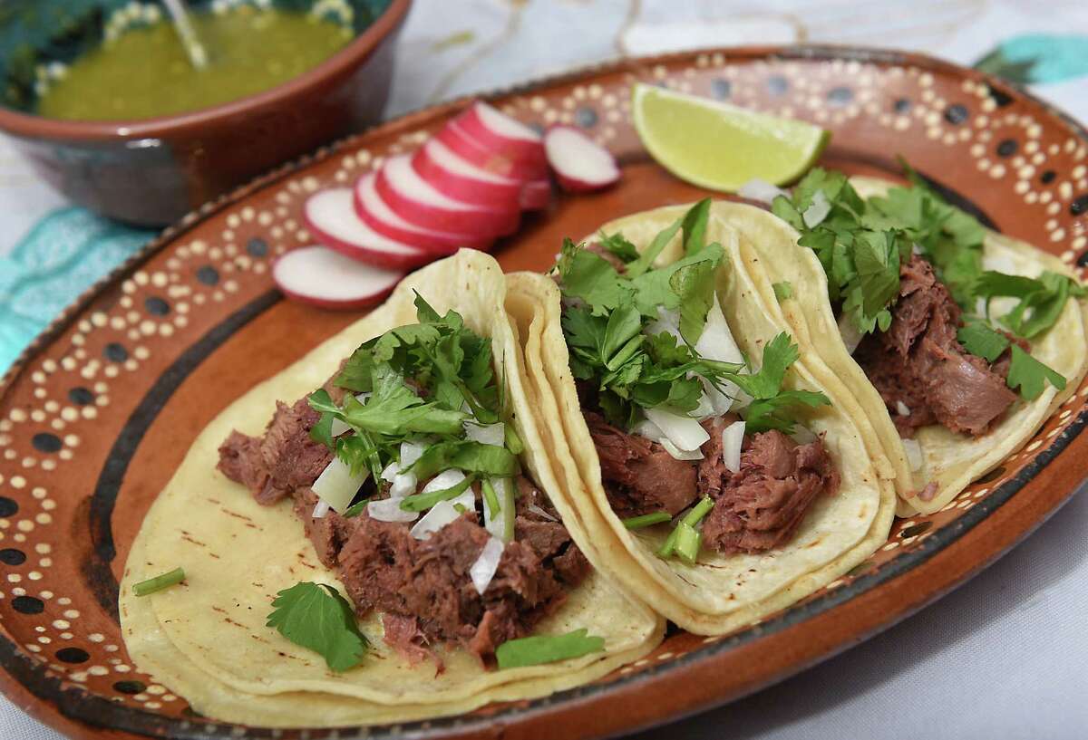 Three cow tongue (lengua) tacos topped with onion and cilantro sit on a brown decorative oval plate. There is sliced radish and lime on the plate, and a bowl of salsa in the background.