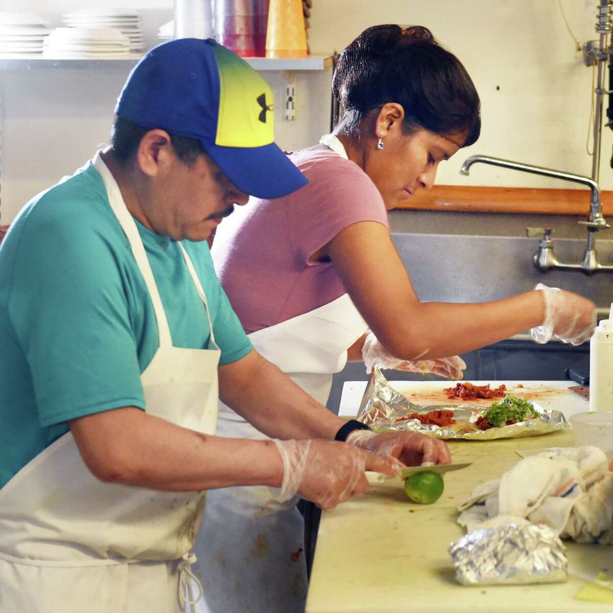 A man in a teal T-shirt, white apron and blue and yellow hat slices limes with gloved hands. A woman next to him in a mauve shirt and white apron tops tortillas with taco fillings.