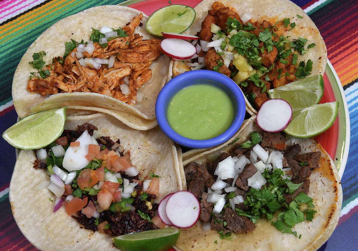 Four tacos with various fillings take up a quarter each of a round plate. Chopped white onions, sliced radish and lime wedges are all over the tacos and the plate. A small round blue dish of green sauce sits in the middle. The plate sits on a striped surface.