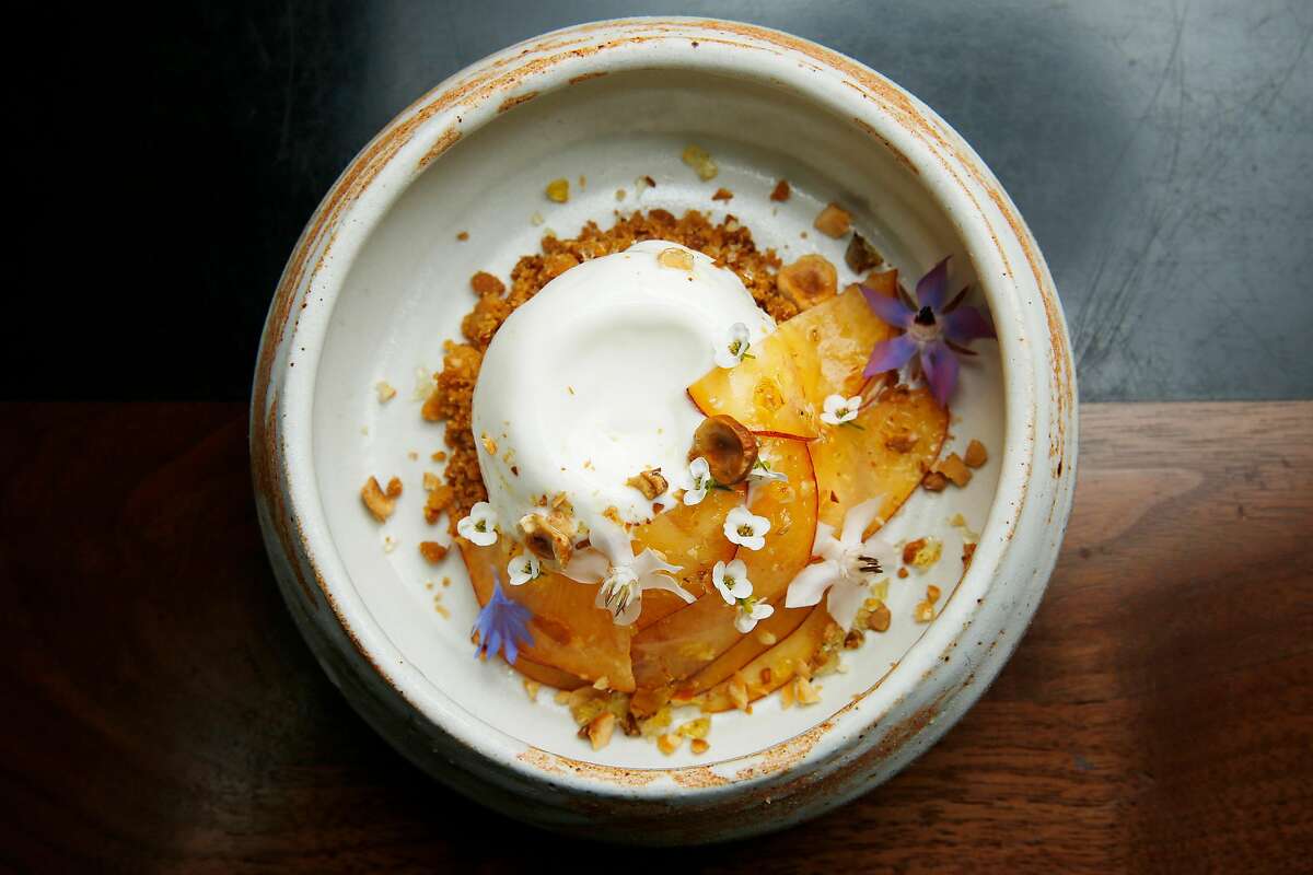 A frozen white dessert topped with shaved stone fruit, nuts and edible flowers.