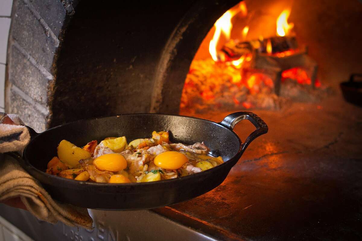 A chef puts a cast iron pan filled with hash and eggs into a wood-fired oven.
