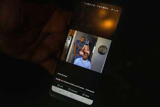A hand holds a phone with a Facebook image of Anthony and two others. One person, holding Anthony’s arm, is standing with him behind an armchair in which another person is seated. There are nine comments on the photograph and the icons indicate people reacted with “love,” “like” and “care.”