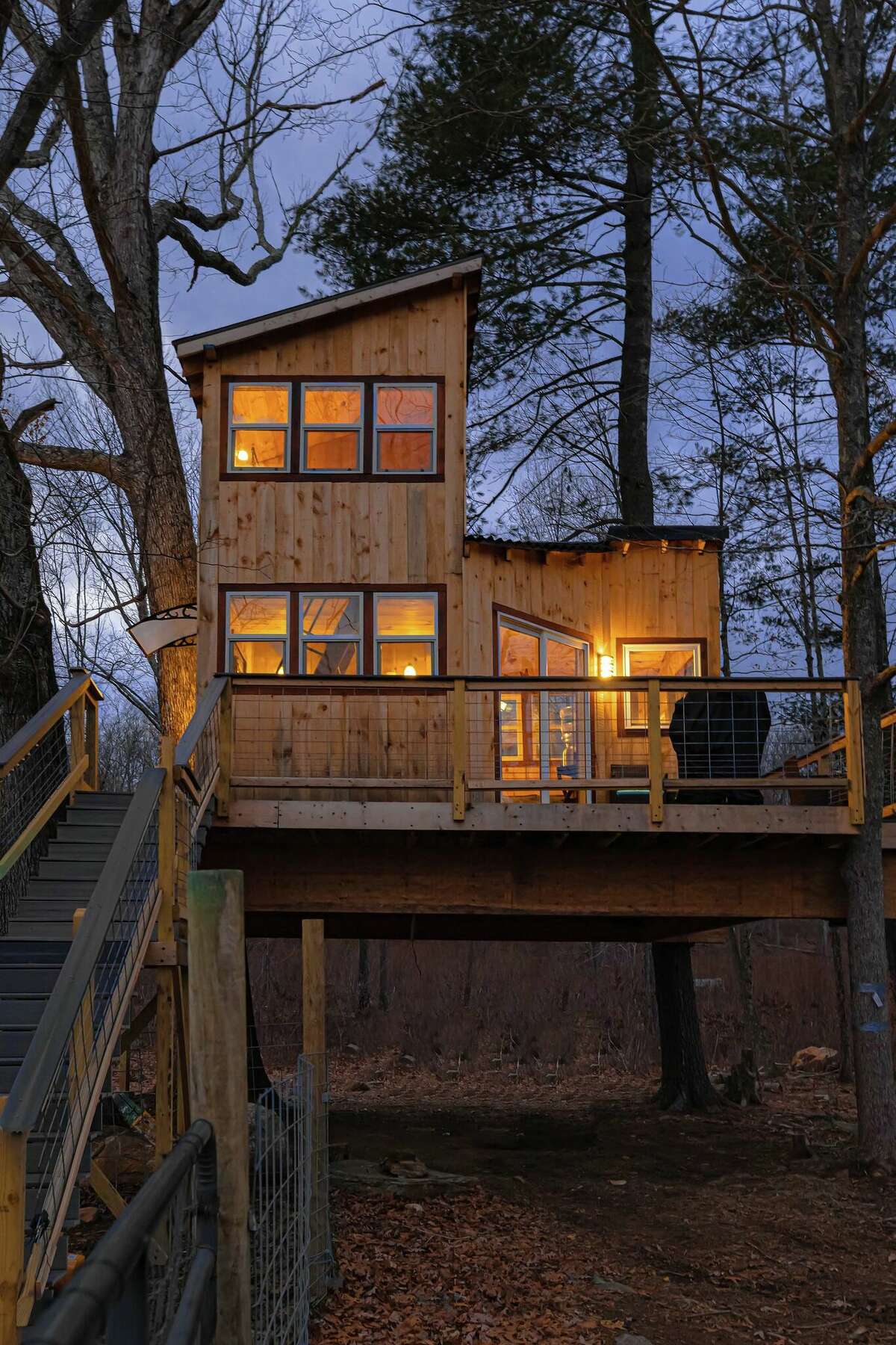 Stairs lead up to a two-story wood cabin sitting on a wooden platform, surrounded by trees. It is nearly sundown and lights are on inside the cabin and by the front door.