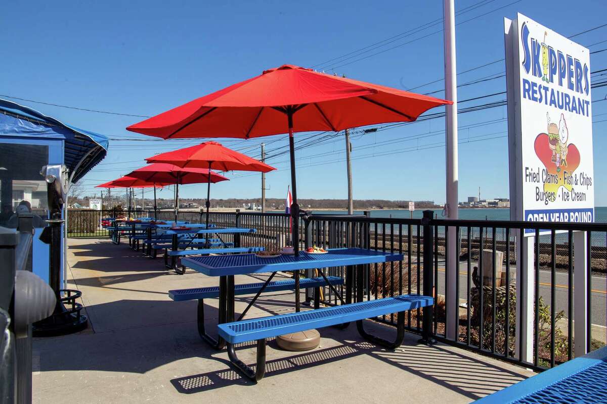 A row of blue picnic tables with red umbrellas on an outdoor patio. Water is visible in the distance.