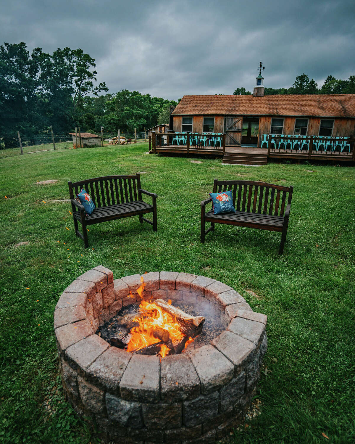 A stone firepit with an open fire two wooden benches. Behind that is a wood cabin with a long deck and an animal pen, with trees in the background.