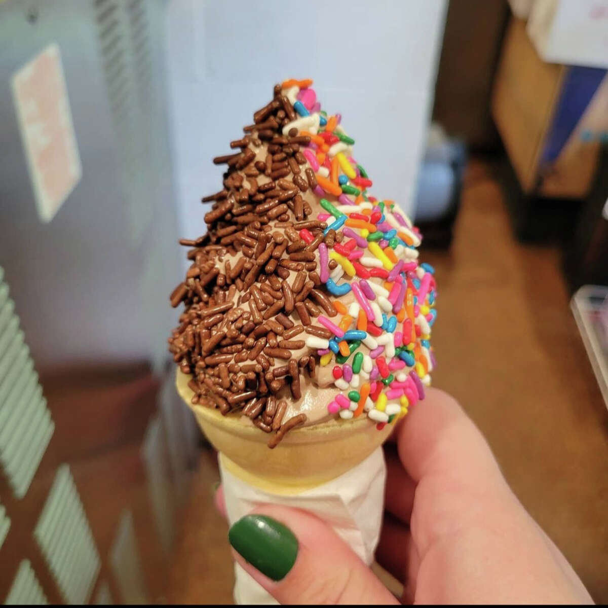 A hand is holding an ice cream cone. The ice cream is vanilla and is covered with chocolate sprinkles on the left side and rainbow sprinkles on the right side.
