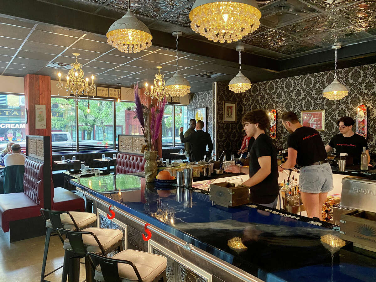 Interior of a restaurant. A blue bar with three stools with white cushions. You can see two maroon booths for seating. You can see seven people: Two employees in black shirts behind the bar, a man in a blak shirt at the far end of the bar, and silhouettes of two pairs of people nearer to the windows facing traffic. There are two chandeliers and five round light fixtures on the ceiling.