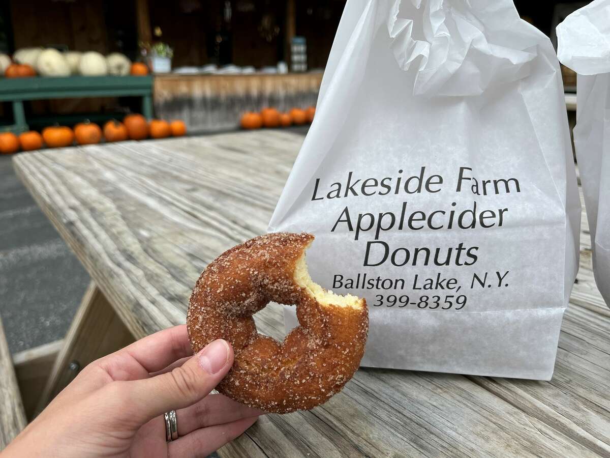 A hand holds a cider doughnut with a bite out of it in front of a white paper bag for Lakeside Farms.