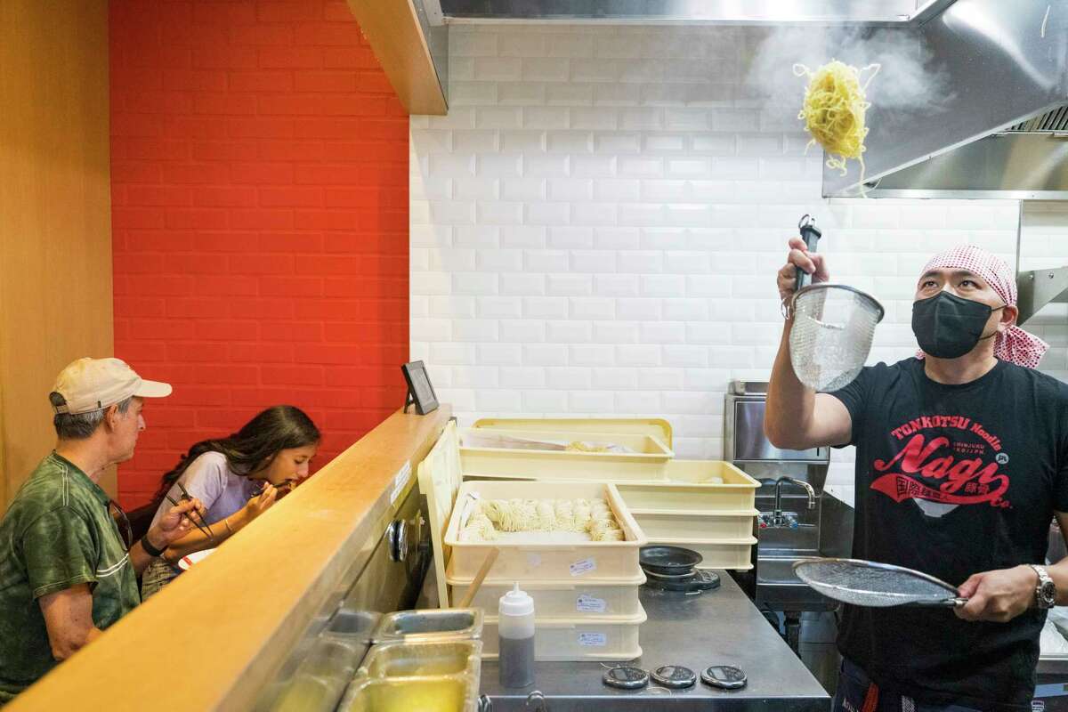 Two people sit at a counter while a chef tosses a bundle of ramen noodles in the air.