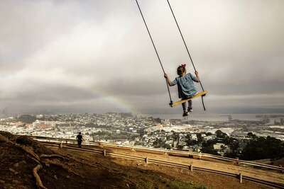 A girl flies high on a tree swing at Bernal Heights Park while, in the background, a rainbow appears over a vista of San Francisco.