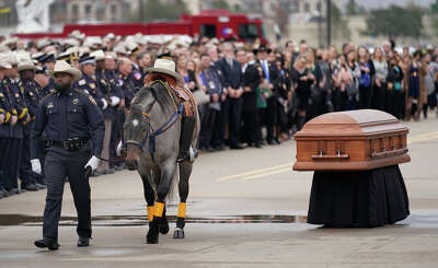 A casket sits on a stand as a horse with no one riding on it but a cowboy hat in the saddle is led away by a police officer.