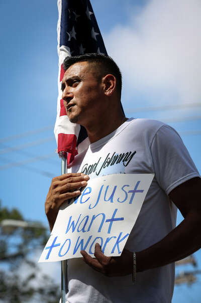 A man stands holding a sign that says "we just want to work" while holding an american flag against his shoulder with a sullen expression on his face against a blue sky