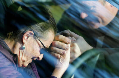A woman bows her head in prayer as she locks hands with a priest through the window of a car as seen through reflections on the windshield of a car.