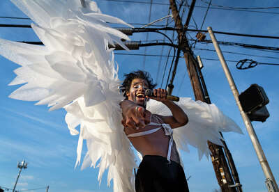 A shirtless man with facepaint and wearing giant white angel wings points at the camera as he holds a microphone and sings in front of a blue sky and powerlines 