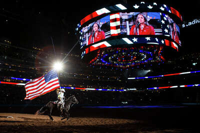 A flare from a light glints off of the top of an American Flag carried by a galloping horse rider as she is spotlit running through a dark stadium with a jumbotron showing the woman singing the National Anthem.
