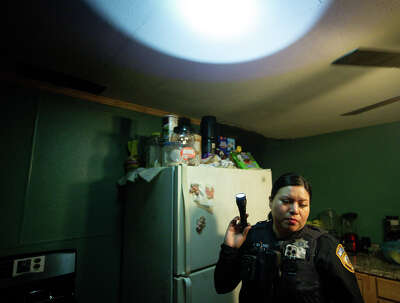 A police officer stands next to a refridgerator in a dark kitchen, her flashlight illuminating the ceiling as she looks away.