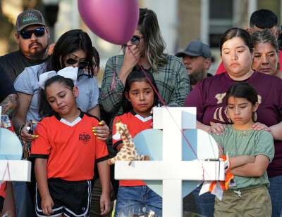 A white cross with a stuffed animal sits in the foreground as three young girls look sad as they look at the memorial made up of crosses.