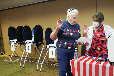 Two women raise their right hands to pledge in oath in a room with voting machins behind them and a table in front of them covered by an American Flag table cloth