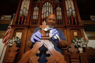 A man wearing gloves uses a rag to wipe down a piece of wood in a church in front of a large organ