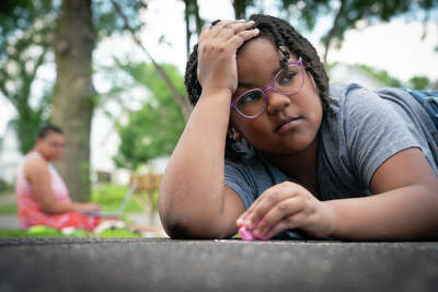 A young firl with braids and glasses looks to the side as she holds one hand to her head and one holds a piece of chald she is laying on the driveway and drawing with