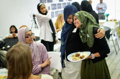 Two women share a smiling embrace as they hold plates of food and talk to a table of friends in a room.