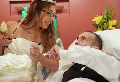 A woman in a wedding dress smiles at a man in a tuxedo laying in a hospital bed dressed in a tuxedo as they dab cake on each other's noses.