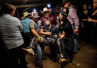 Three men carry a man who is wearing a helmet and leg pads and appears unconcious away from a rodeo ring 