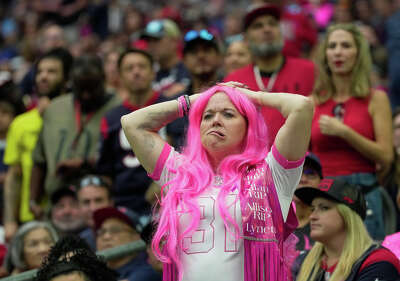 With a crowd of fans behind her, a woman wearing a pink wig stands and puts her hand on her head with an exasperated expression..