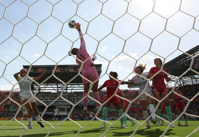A soccer goal net fills the foreground through which you see a goalie jumping and reaching into the air to barely get her hand on the ball over a group of onlooking soccer players.