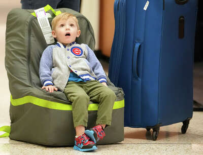 A young boy wearing a Chicago Cubs sweater and tennis shoes sits back on a piece of luggage, looking up with a bemused look on his face.
