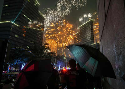 Fireworks explode in the sky around two buildings in front of a crowd of people holding umbrellas