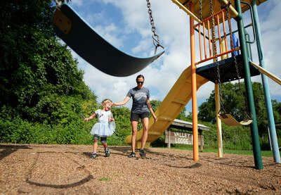 A woman walks with her daughter through a playground surrounded by blue sky and greener while a swing hangs in the foreground of the photo
