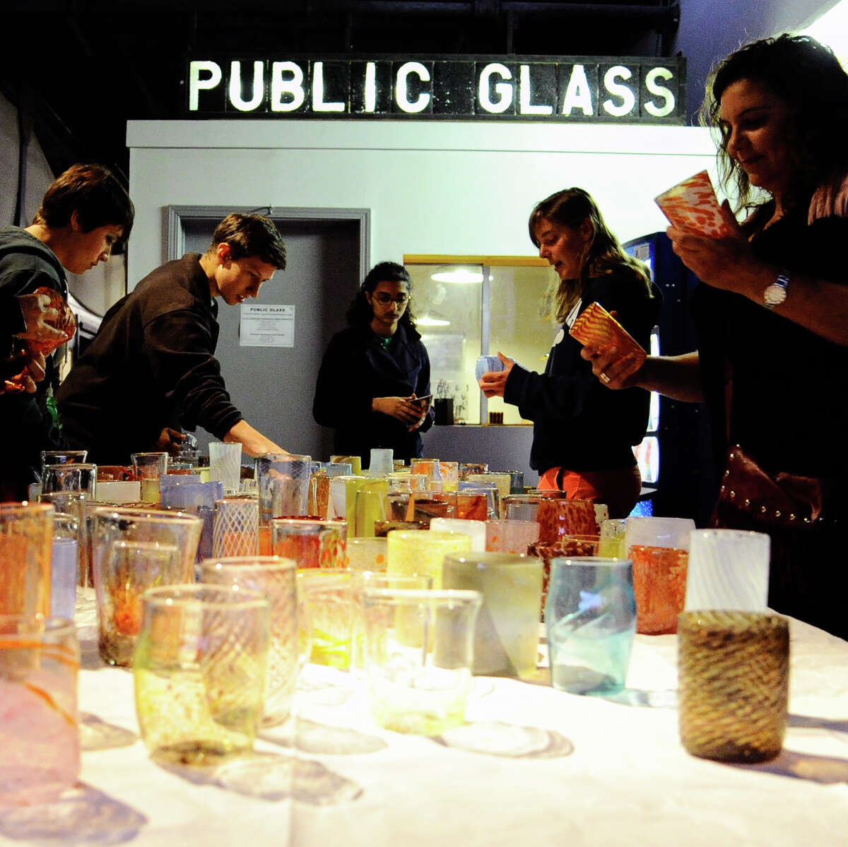 A table holds glass art that is given away at the Hot Glass Cold Beer event. A 'Public Glass' sign is over the table and people are milling around