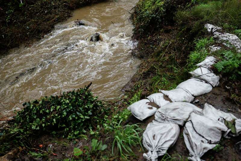 A group of sandbags line a creek with brown rushing water.