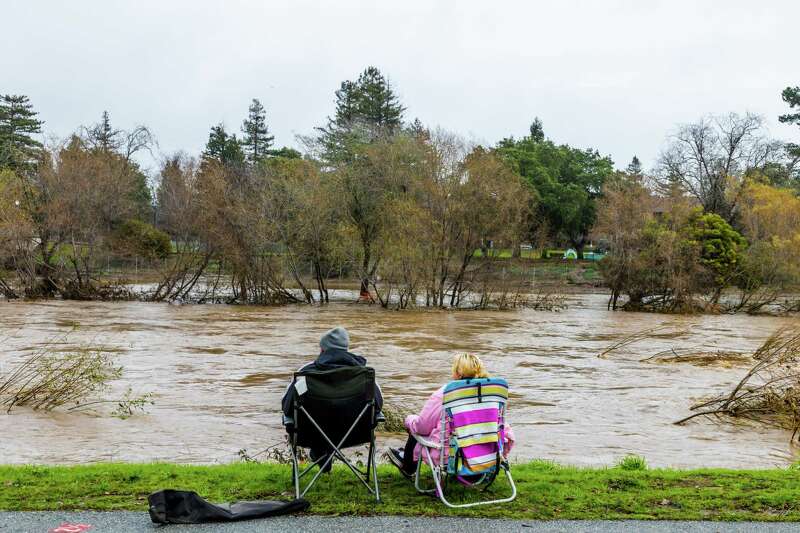 Two people sit on lawn chairs on the banks of a flooded San Lorenzo River.