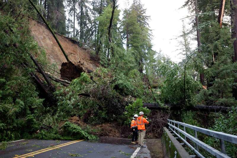 Two people wearing orange vests study trees and other debris blocking a roadway in a forested area.
