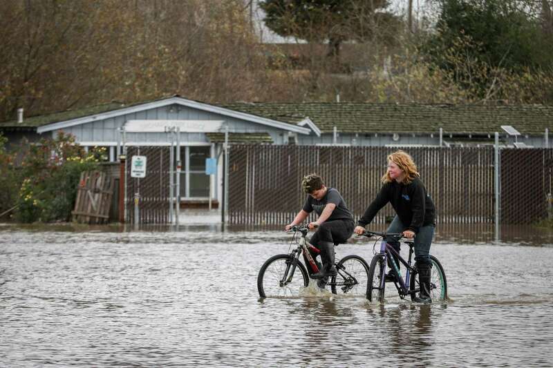 Two kids ride their bikes along a flooded street.