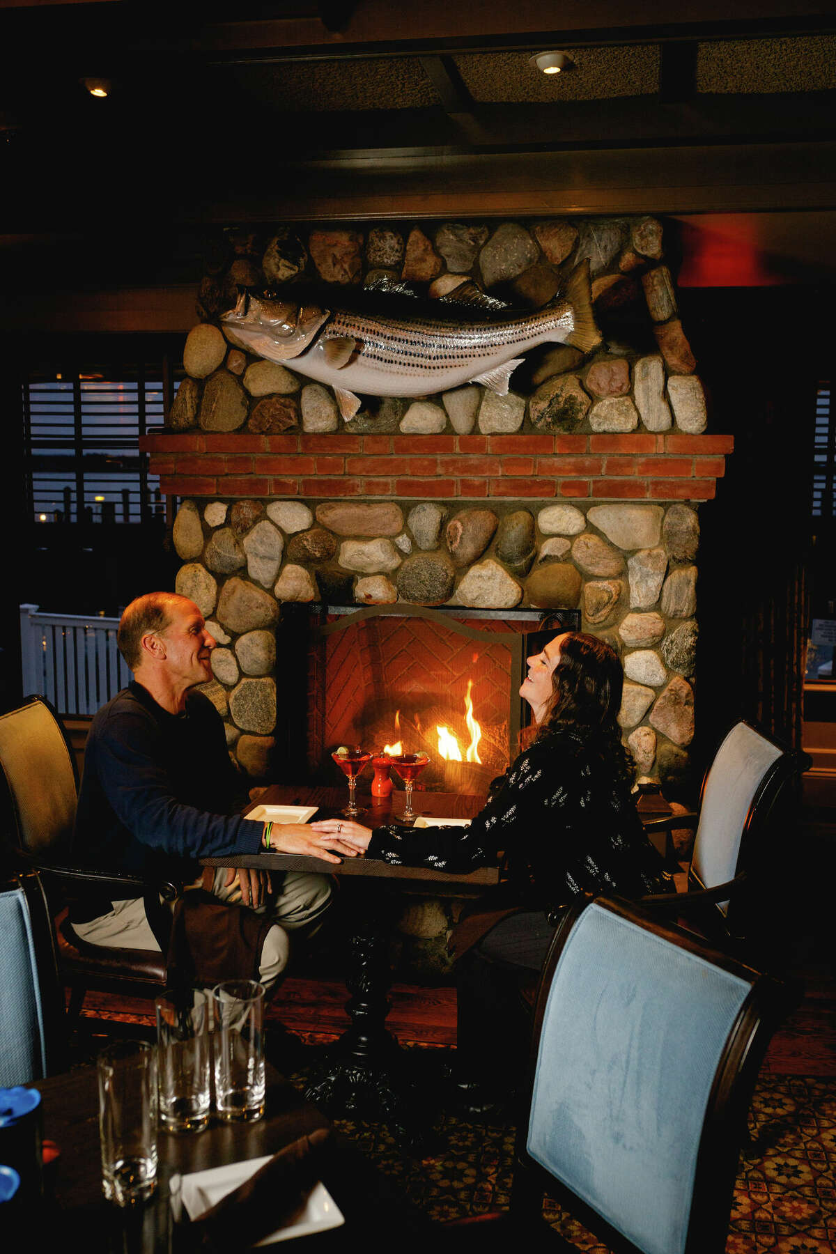 A couple eating and laughing in front of a lit stone fireplace in a cozy restaurant setting