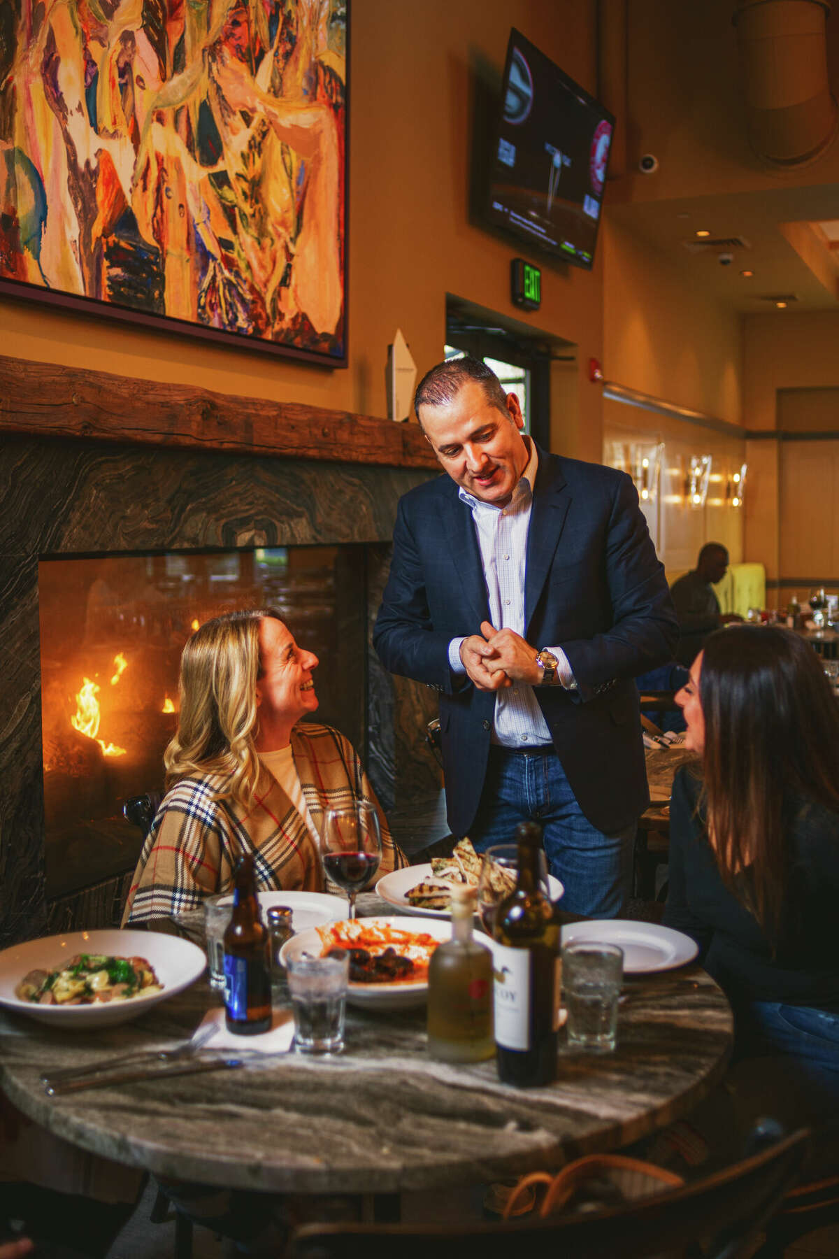 a host speaks with two women having dinner in front of a roaring fireplace