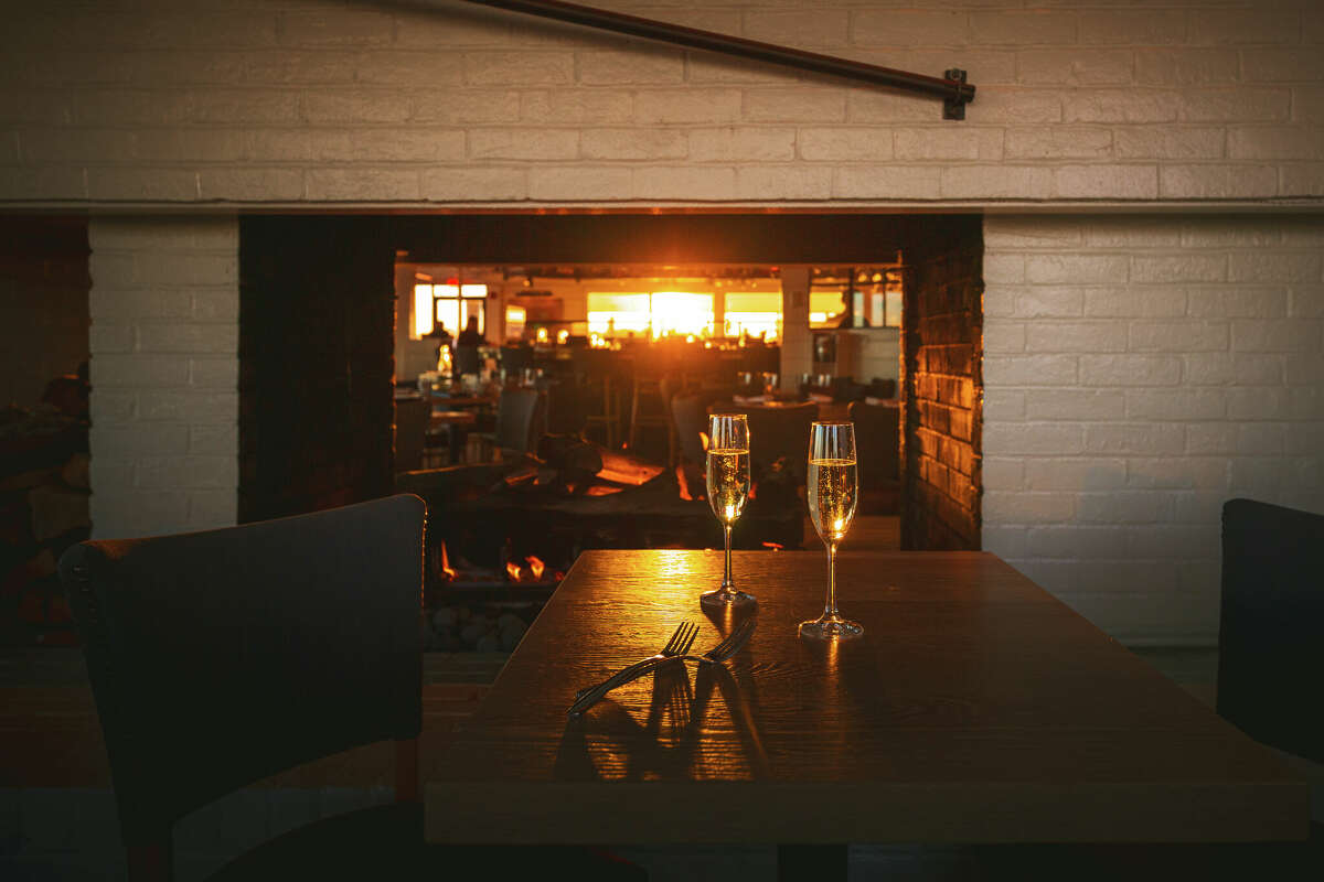 a dimly lit restaurant interior, with two champagne flutes on a table int he foreground and the sun setting through the window in the background.