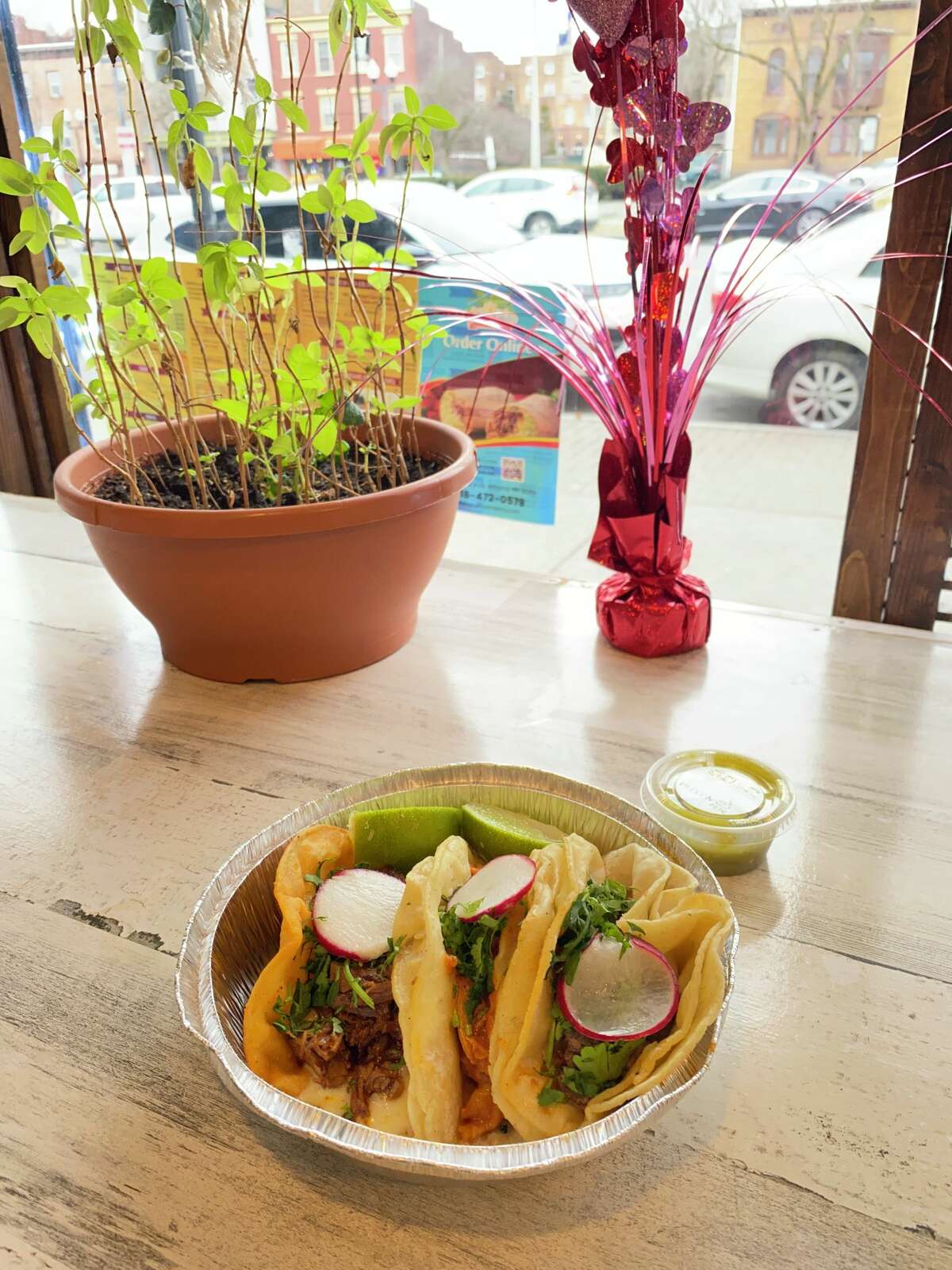 Three tacos in a tin foil tray sit on a wood table. A green plant and pink decoration can be seen in front of the window, looking out toward traffic.