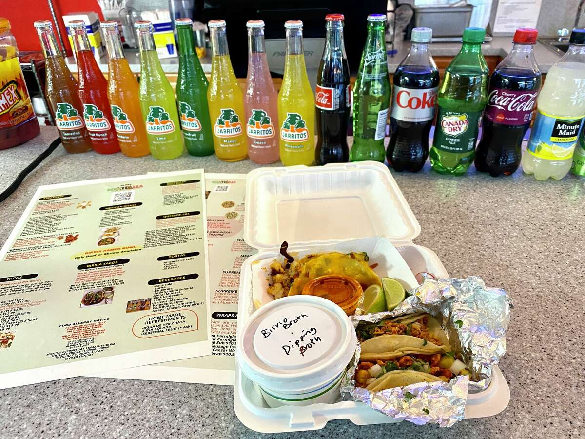 A styrofoam tray with two comparments holds a taco in the background with a red salsa and sliced limes. In the foreground, two meat-filled tacos with onion and cilantro are wrapped in partially opened foil, and sit next to a cup of birria broth. Menus can be seen on the left, and in the background are glass and plastic bottles of sodas and drinks.