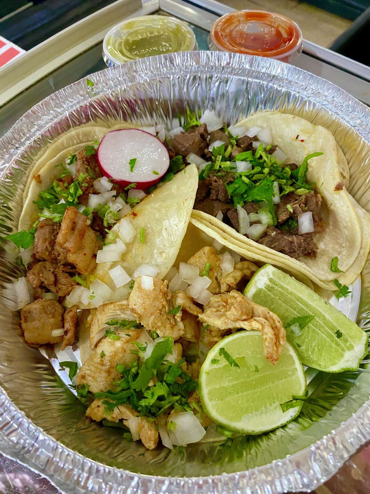 Two meat-filled tacos topped with radish and onions sits in a foil container, with sides of sliced limes and a side dish