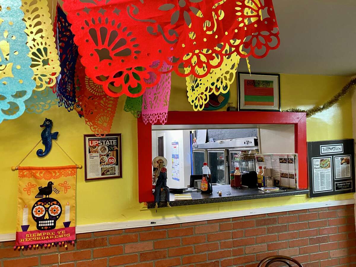 A grocery service counter window sits in a yellow and brick wall. There are hangings on the ceiling in red, pink, yellow and blue. Framed art surrounds the service window.