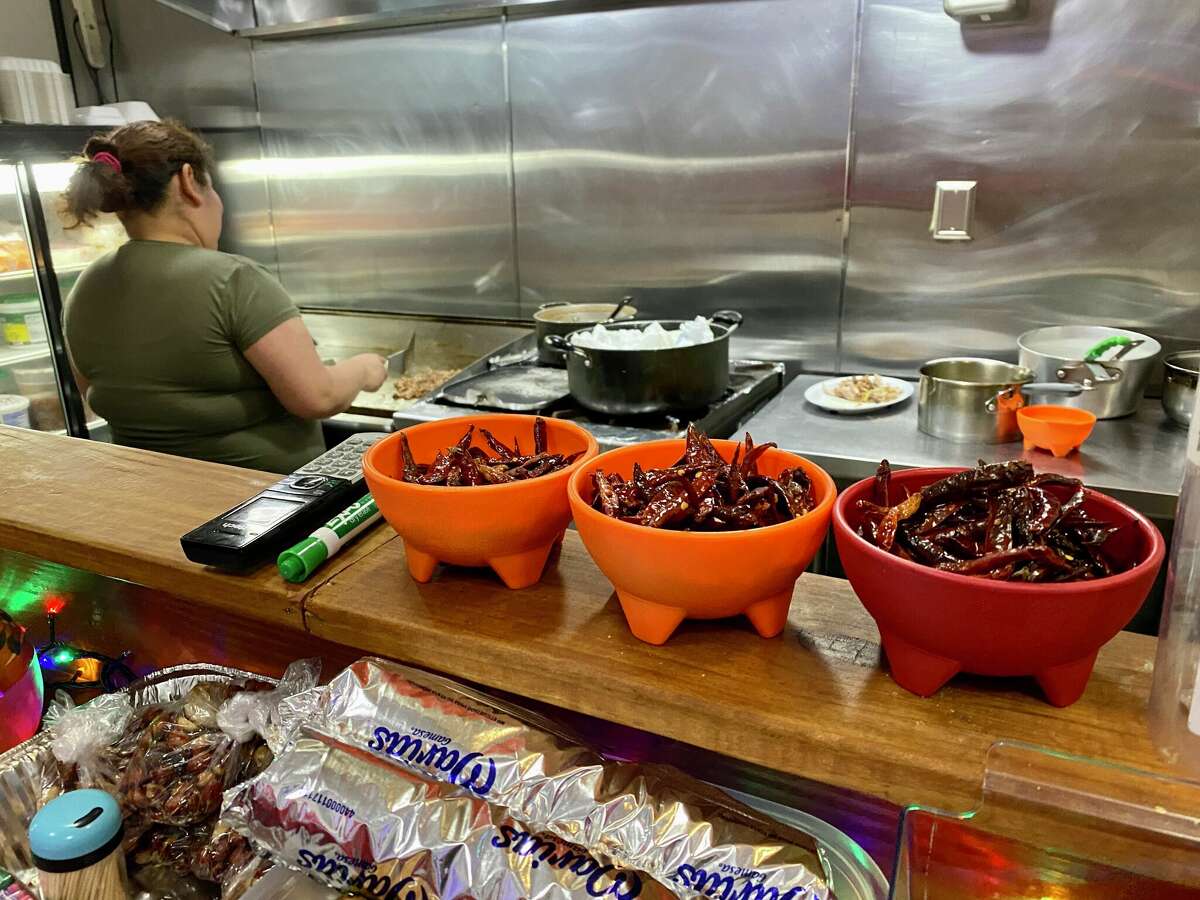 A service counter of a restaurant. Three bowls of dried chiles are in the foreground. You can see a person in an olive green shirt tending to ingredients in a stainless steel kitchen.