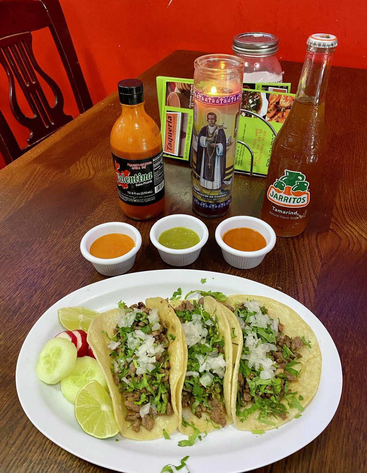 In the foreground, three tacos with meat, onion and cilantro sit on a white plate with sliced limes, cucumber and radishes. Behind the plate are three white dishes of sauces, a jarritos soda, a bottle of Salsa Valentina and a prayer candle. A menu and a jar can be seen in the background.
