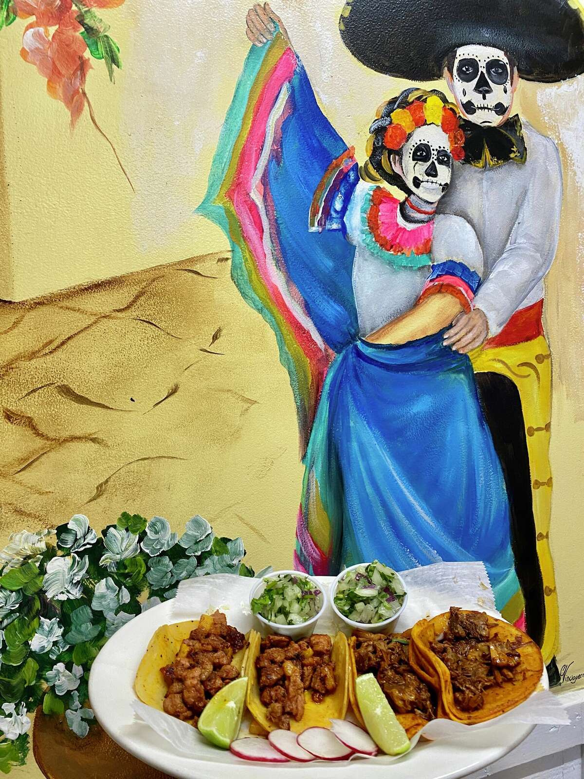In the background, a mural of a man in a calavera/sugar skull mask in yellow pants with a red cumberbund, a white shirt and a black hat and a woman in a blue dress with white, pink and yellow trim, also in a calavera/sugar skull mask and flowers in her hair. They stand at a yellow wall in the painting. In the foreground, four meat filled tacos sit on a white plate with lime wedges and sliced radishes. There are two small cups of a green salad.