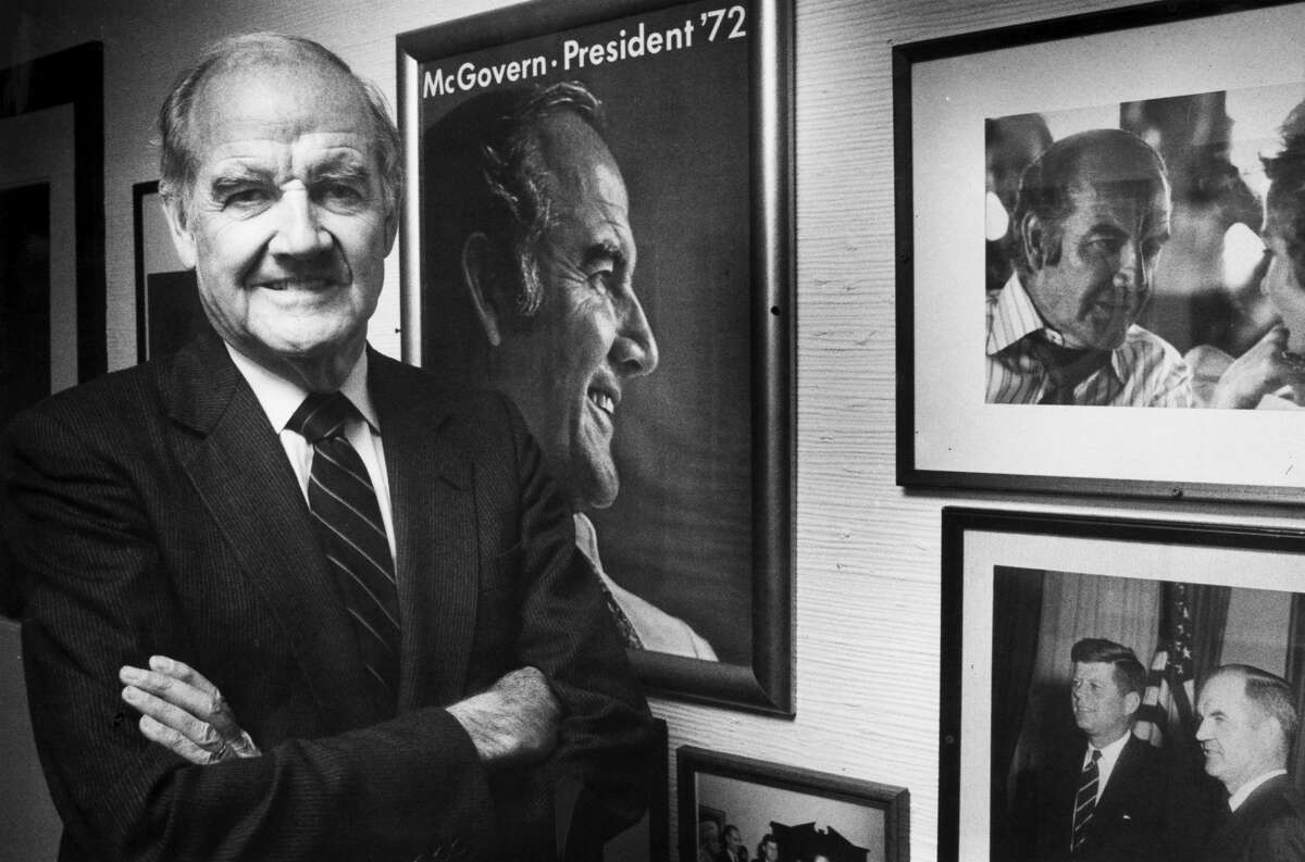 George McGovern, left, poses with an old campaign posters and photographs from his political career, which line a hallway at Stratford Inn in Stratford, Conn., Nov. 12, 1989.