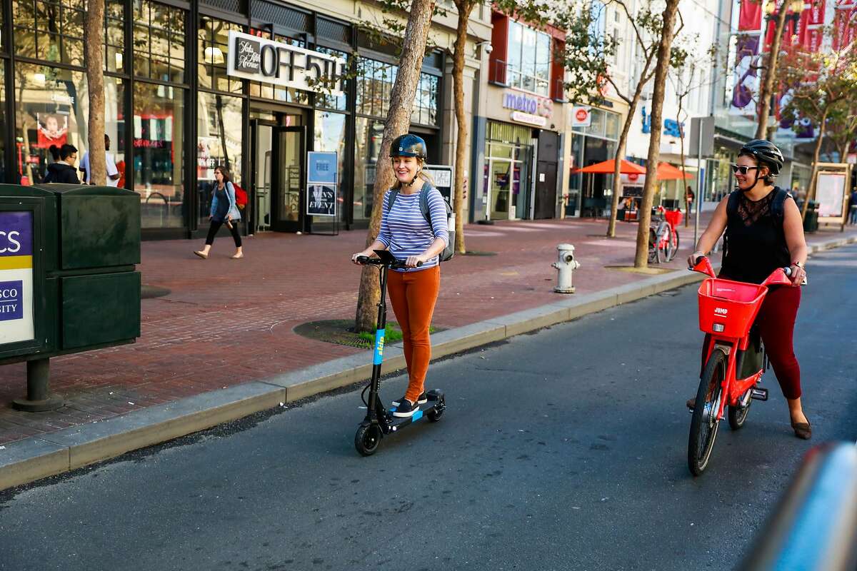A woman rides a Skip scooter while another woman rides an electric bicycle on Market and 5th Streets in San Francisco, California, on Monday, Oct. 15, 2018.